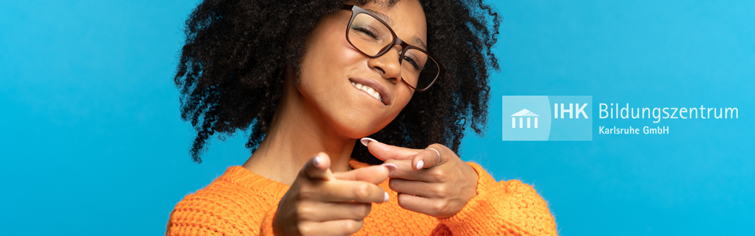 Eine coole junge Frau mit Brille und orangefarbener Kleidung schaut und zeigt selbstbewusst in die Kamera.