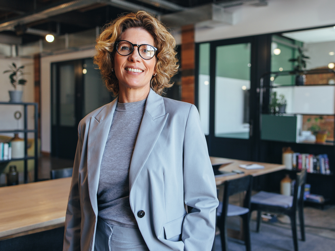 Smiling professional woman in a light gray blazer and glasses stands in a modern open-office setting.