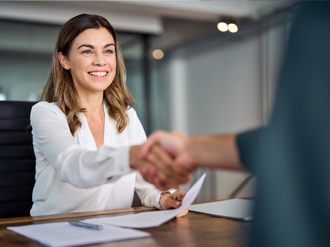 Woman smiling while shaking hands across a desk during a business meeting, with papers on the table.