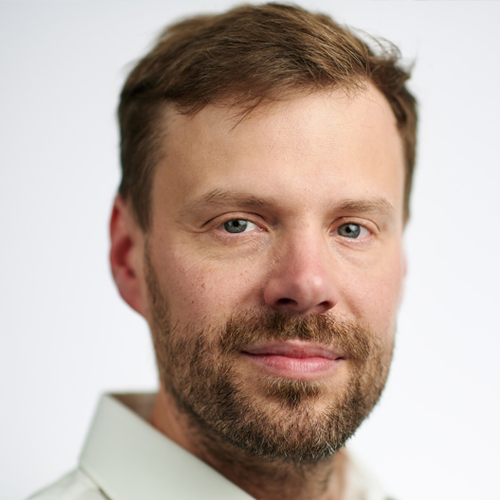 Close-up portrait of a man with short brown hair and a beard, looking at the camera against a light background.