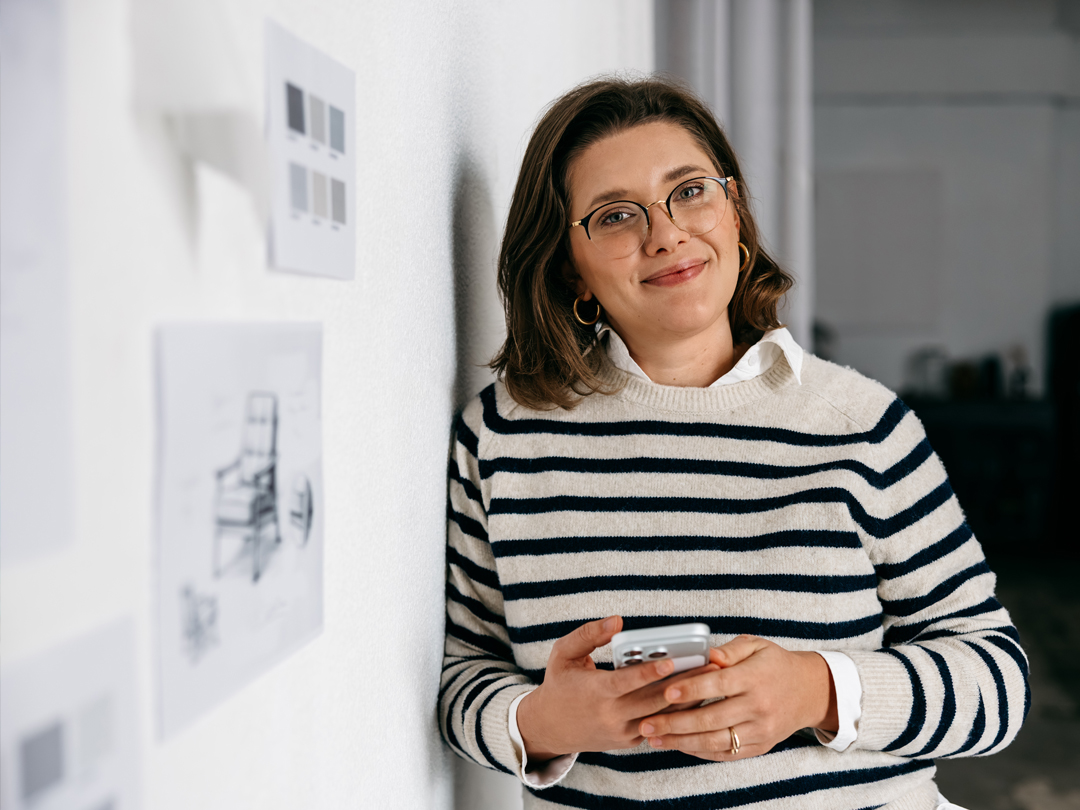 Woman with striped sweater and glasses smiles while holding a smartphone indoors, leaning against a wall with sketches pinned nearby.