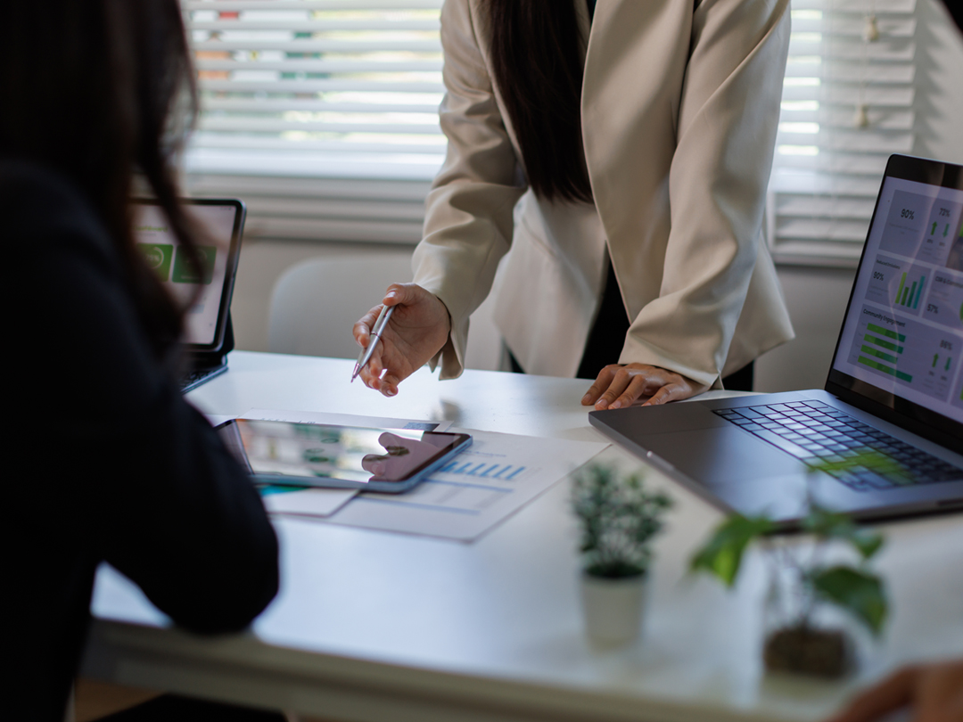 Two professionals discuss analytics at a bright desk: one person in a beige blazer leans over with a pen, a laptop shows charts, and a tablet and papers lie on the table.